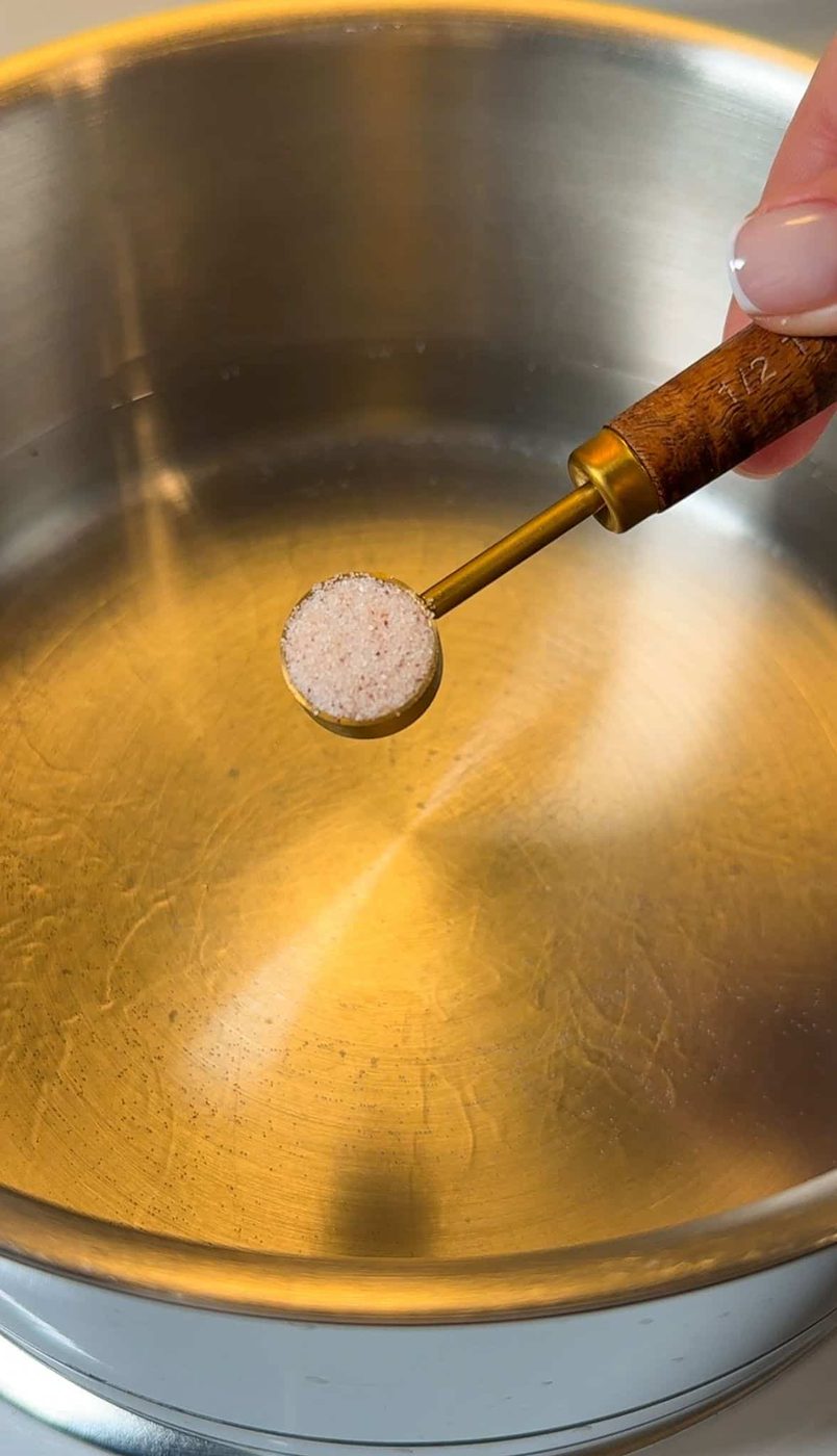 A hand holding a measuring spoon with salt hovers over a stainless steel pot. The pot's interior has a slight sheen, reflecting light, and the scene suggests preparation for cooking.