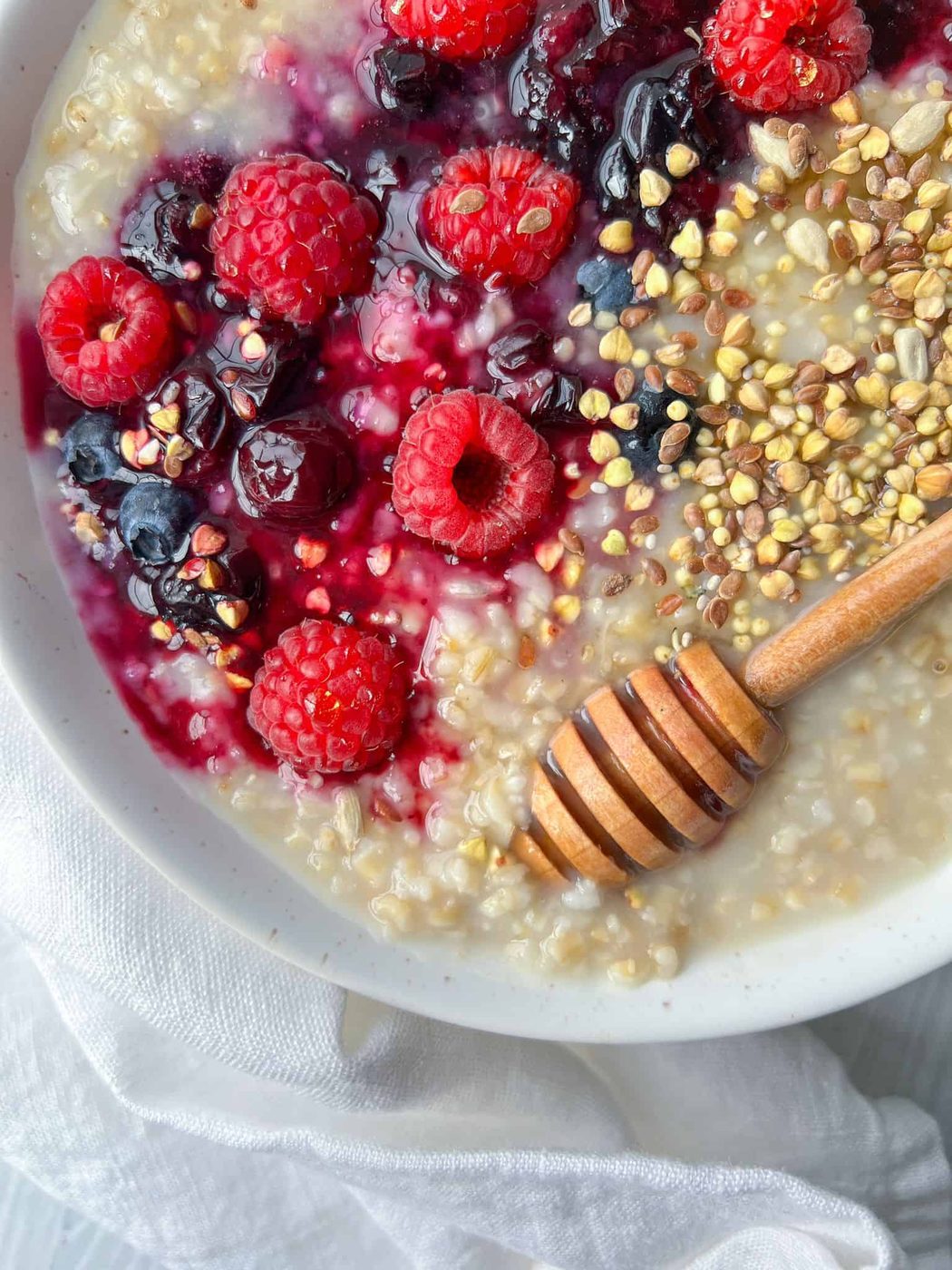 A bowl of steel cut overnight oatmeal topped with fresh raspberries, blueberries, and dark cherries, sprinkled with seeds and nuts. A honey dipper rests on the edge with droplets of honey. The oatmeal is set on a light-colored cloth.