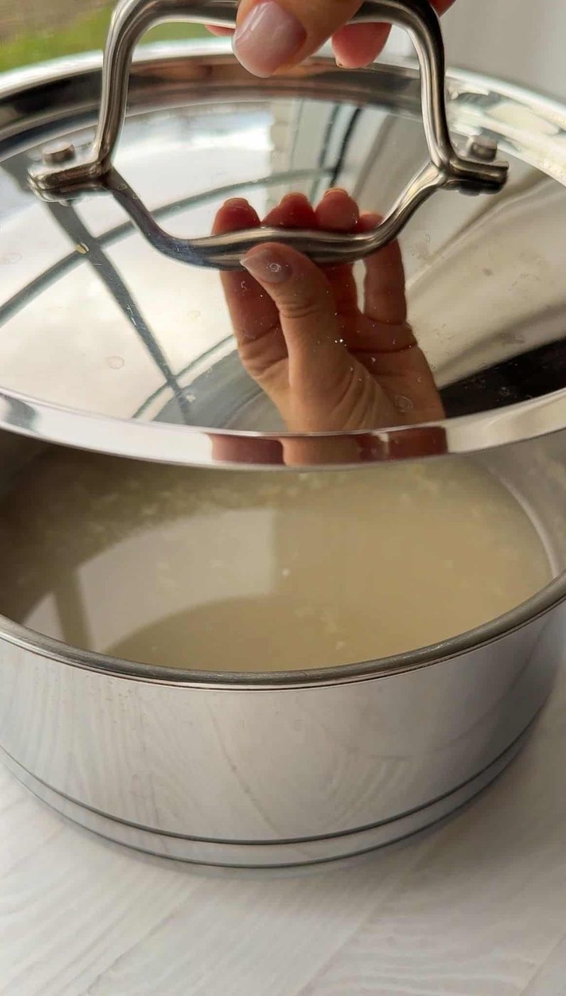 A hand lifts the lid off a metal pot containing a liquid. The pot is on a white surface with natural light streaming in from the background. Water droplets are visible on the lid.