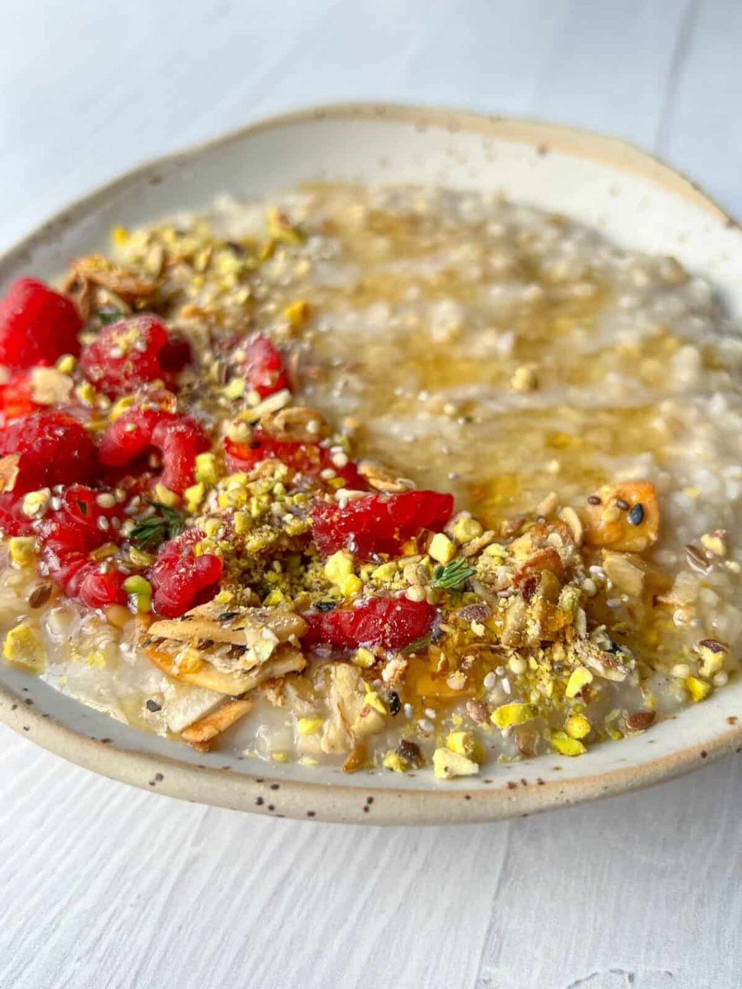 A dish of steel cut overnight oatmeal topped with vibrant toppings, including red raspberries, sliced nuts, and crushed pistachios, drizzled with honey. The oatmeal is served in a rustic, shallow bowl on a light-colored wooden surface.