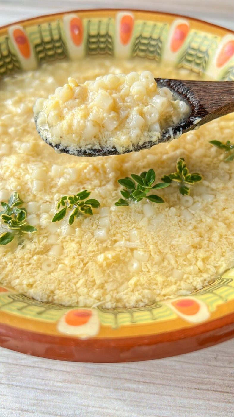 A close-up of a bowl of Stracciatella Soup garnished with green herbs. The soup contains small, round grains and is served in a decorative ceramic bowl. A wooden spoon is lifting a portion of the grains from the soup.