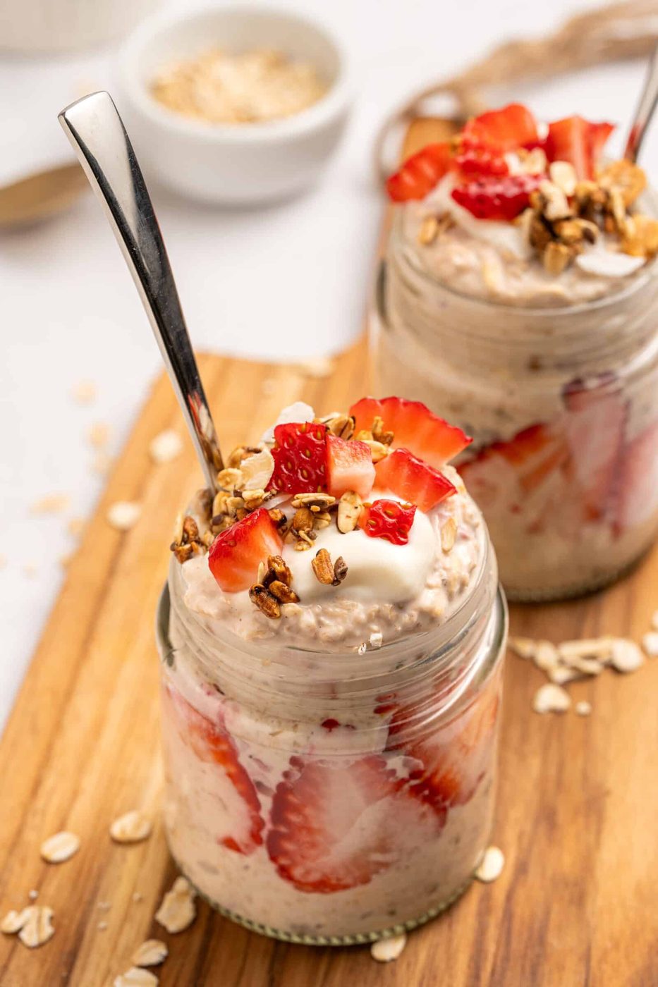 Two glass jars filled, layered with sliced strawberries, topped with yogurt, granola, and fresh strawberries. Each jar has a spoon and sits on a wooden board with oats scattered around.