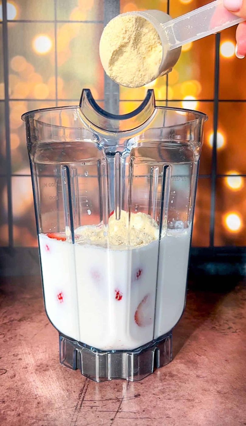 A blender pitcher filled with milk and strawberries on a wooden surface. A hand is adding a scoop of brown powder into the blender. The background features warm, blurred lights creating a cozy ambiance.