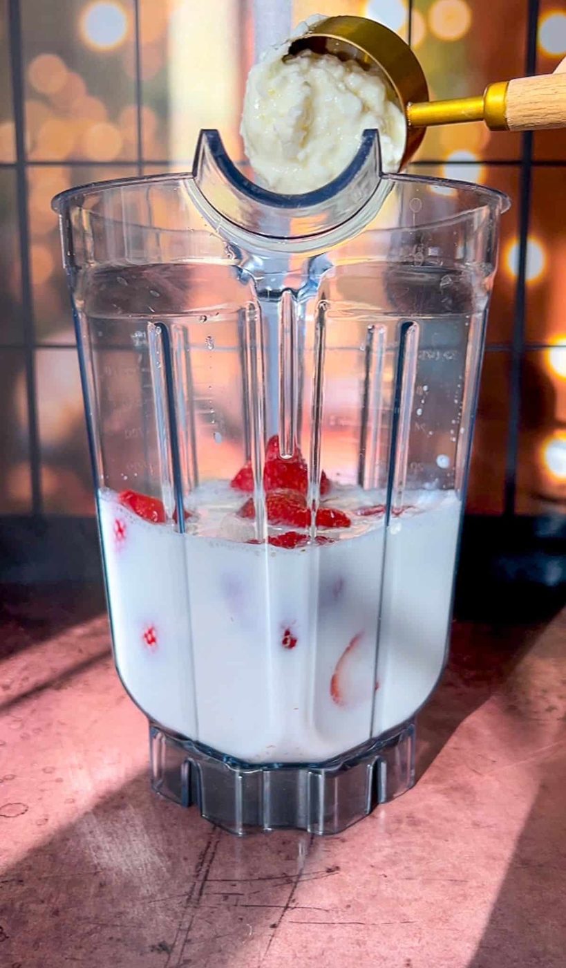 A blender jug filled with milk and strawberry slices sits on a countertop. A golden measuring cup pours a thick mixture, possibly banana puree, into the blender. The background is softly blurred with warm lights.