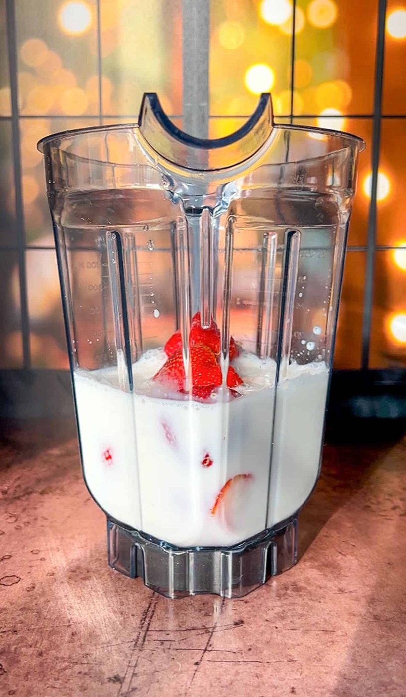 A blender jar filled with milk and sliced strawberries sits on a wooden surface. The background features warm, out-of-focus lights, creating a cozy atmosphere.