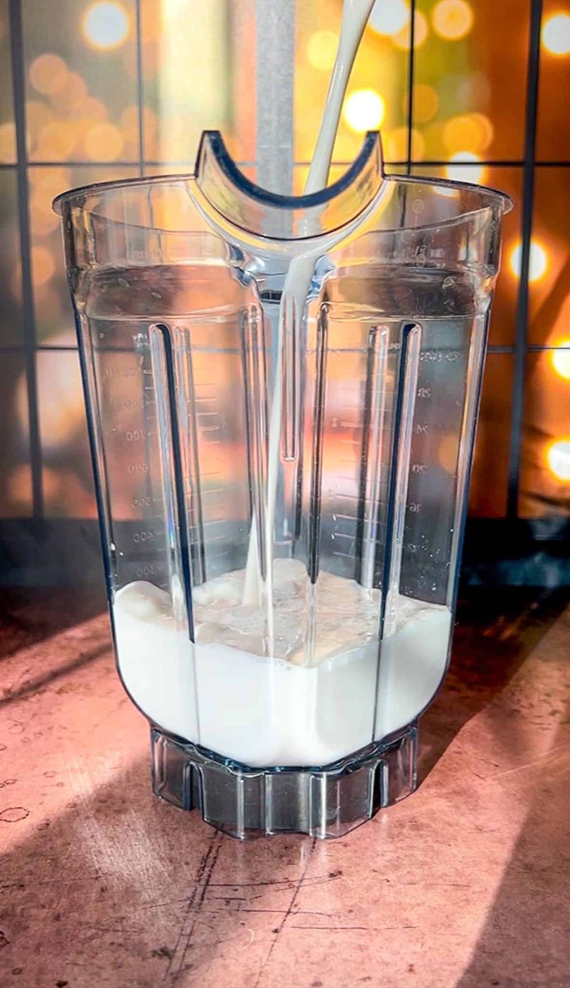 A blender pitcher being filled with milk, set on a wooden surface. The background is blurred with warm, golden bokeh lights, giving the scene a cozy and inviting atmosphere.