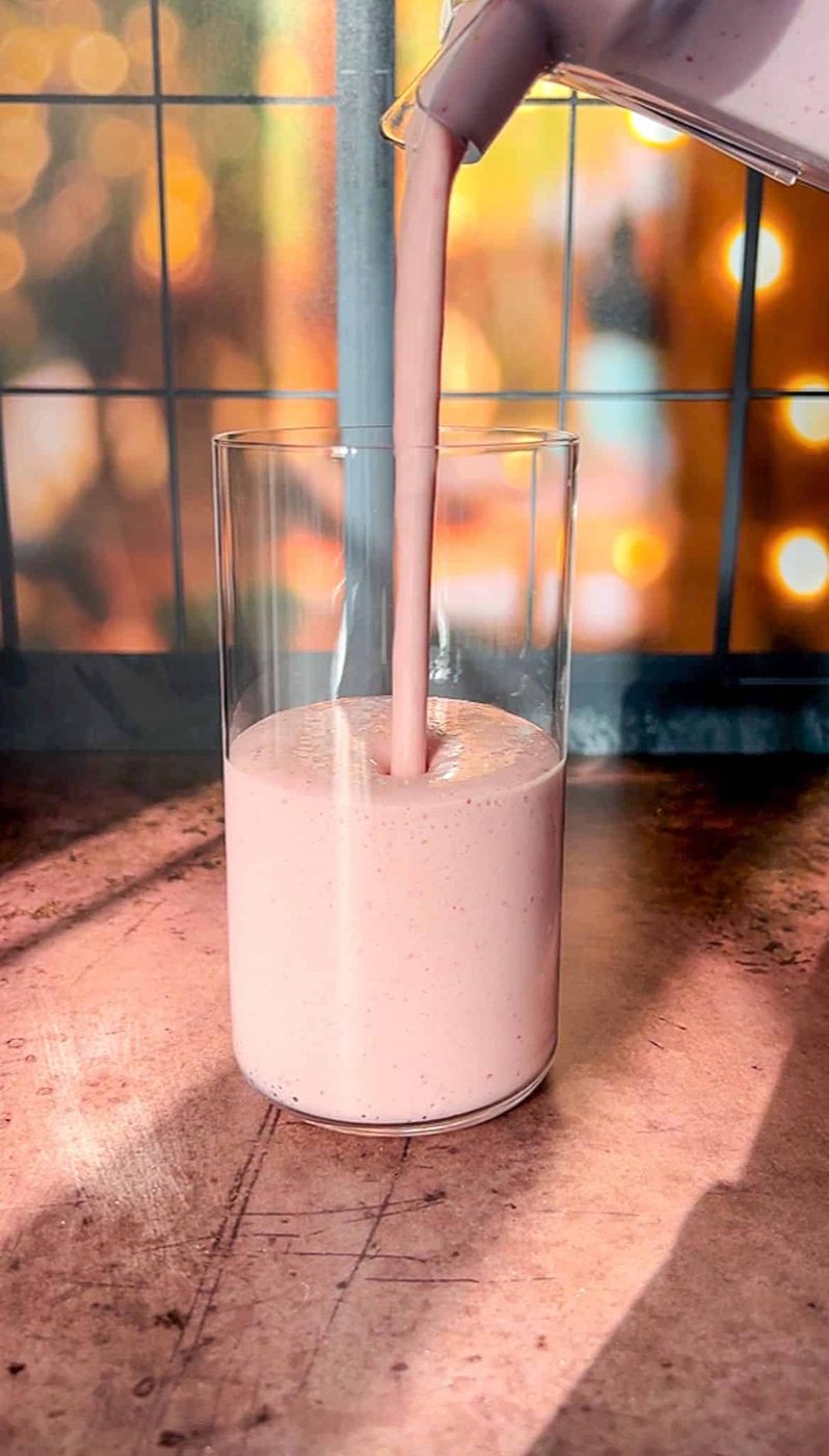 A tall glass on a wooden table is being filled with a strawberry protein shake being poured from a blender. The background has warm bokeh lights and a grid-patterned window, creating a cozy atmosphere.