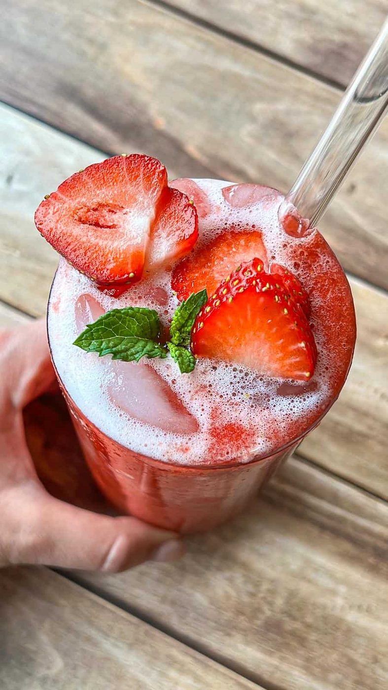 A hand holds a glass of iced strawberry drink topped with foam, fresh strawberry slices, and a mint sprig, with a clear straw, on a wooden surface.