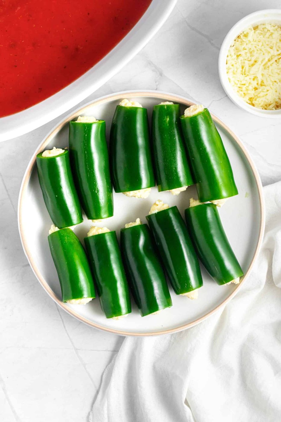 A plate of green jalapenos filled with cheese, ready for baking. A bowl of tomato sauce and grated cheese are nearby on a marble countertop. The peppers are neatly arranged in two rows.