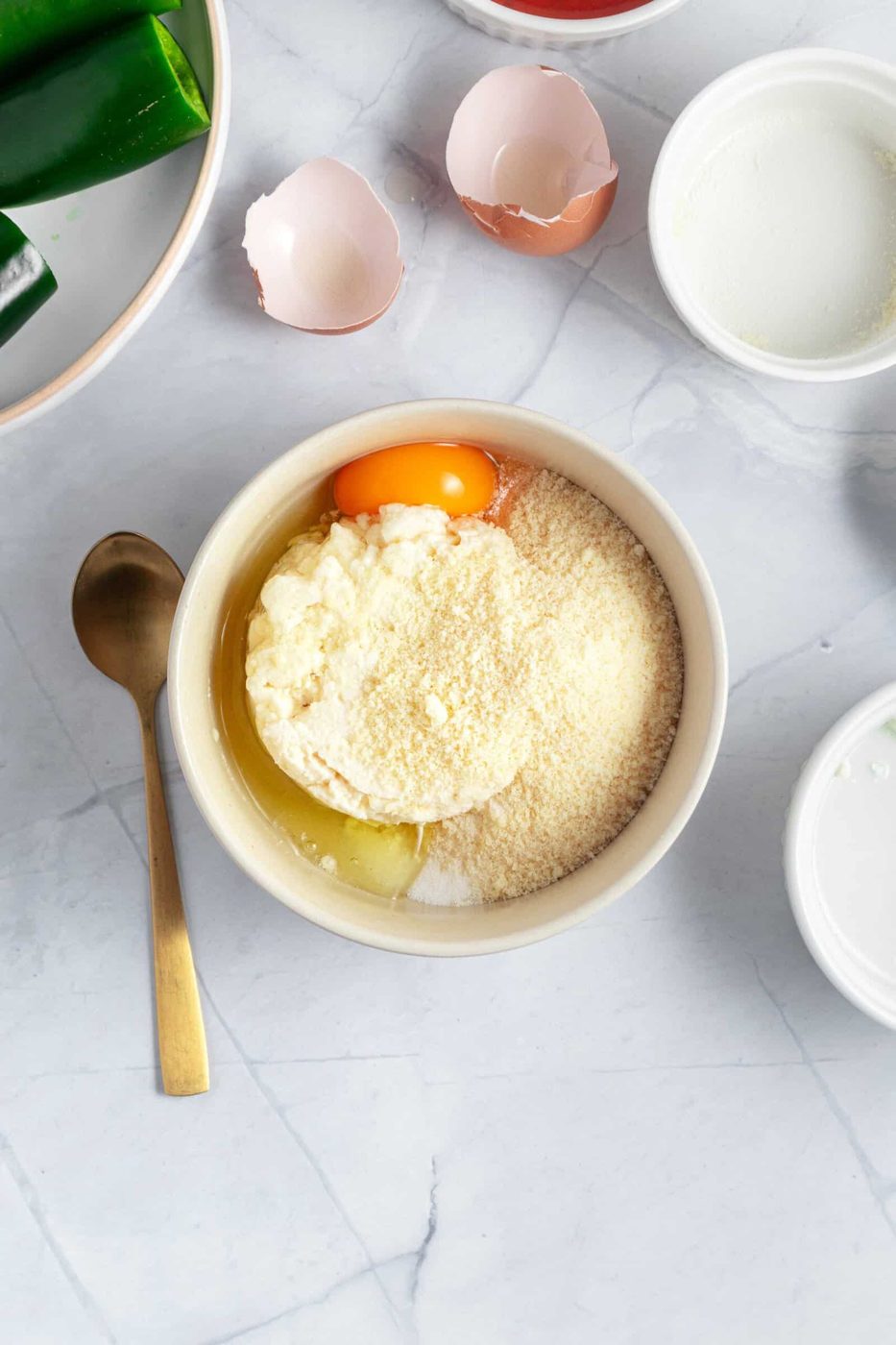 A bowl containing ricotta cheese, grated Parmesan, and an egg yolk on a marble countertop. Surrounding it are broken eggshells, a gold spoon, a white empty bowl, and a plate with green vegetables.