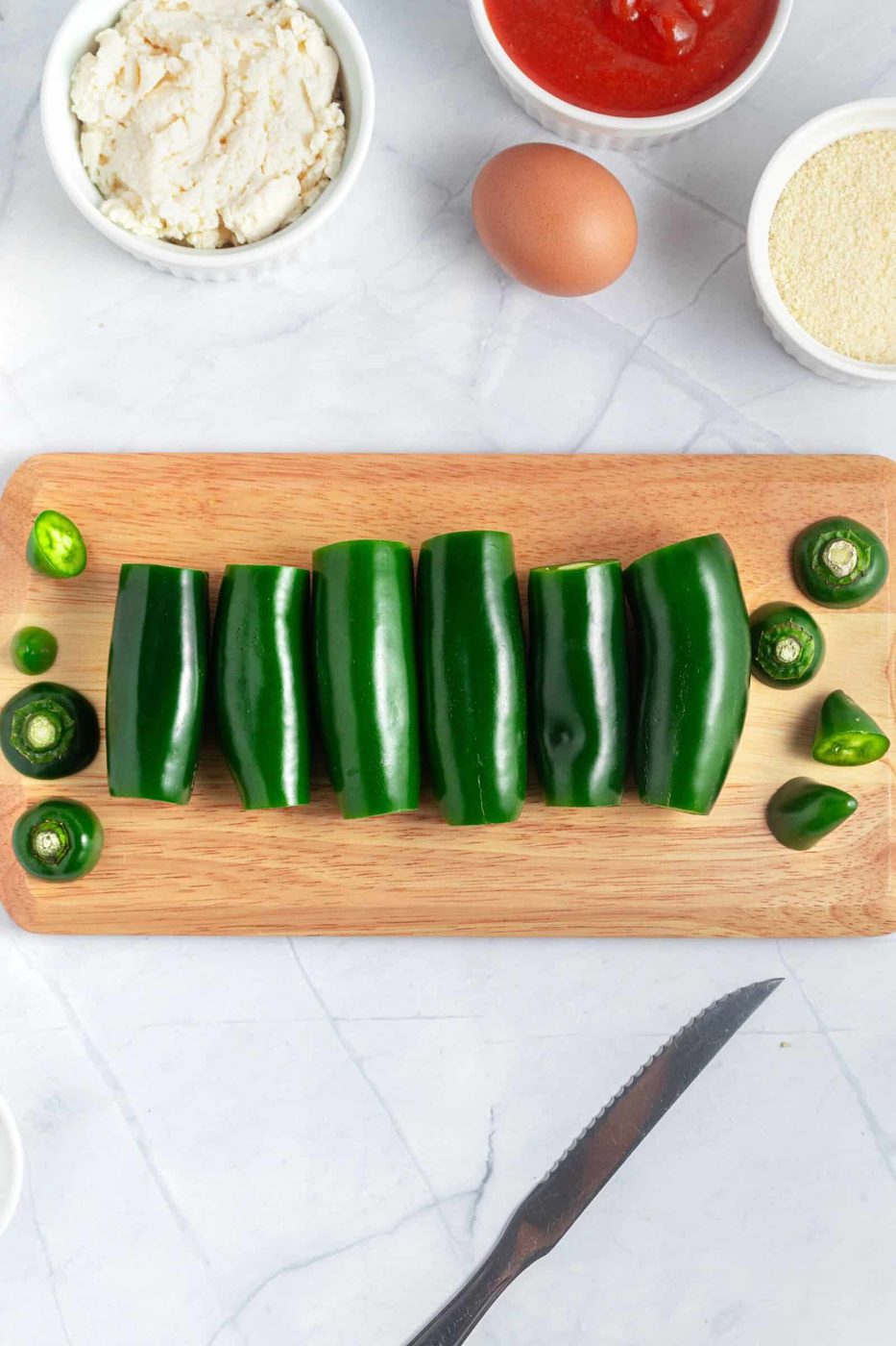 Top view of six green peppers lined up on a wooden cutting board. Accompanied by a knife, and ingredients like ricotta cheese, tomato sauce, and an egg in small bowls nearby, on a marble surface.