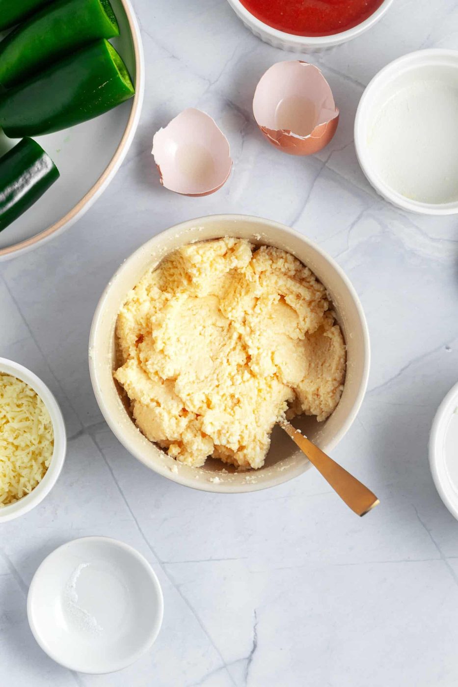 A bowl of creamy mixture with a spoon on a marble surface, surrounded by halved green jalapeños, empty eggshells, a bowl of red sauce, a bowl of grated cheese, and an empty white bowl.