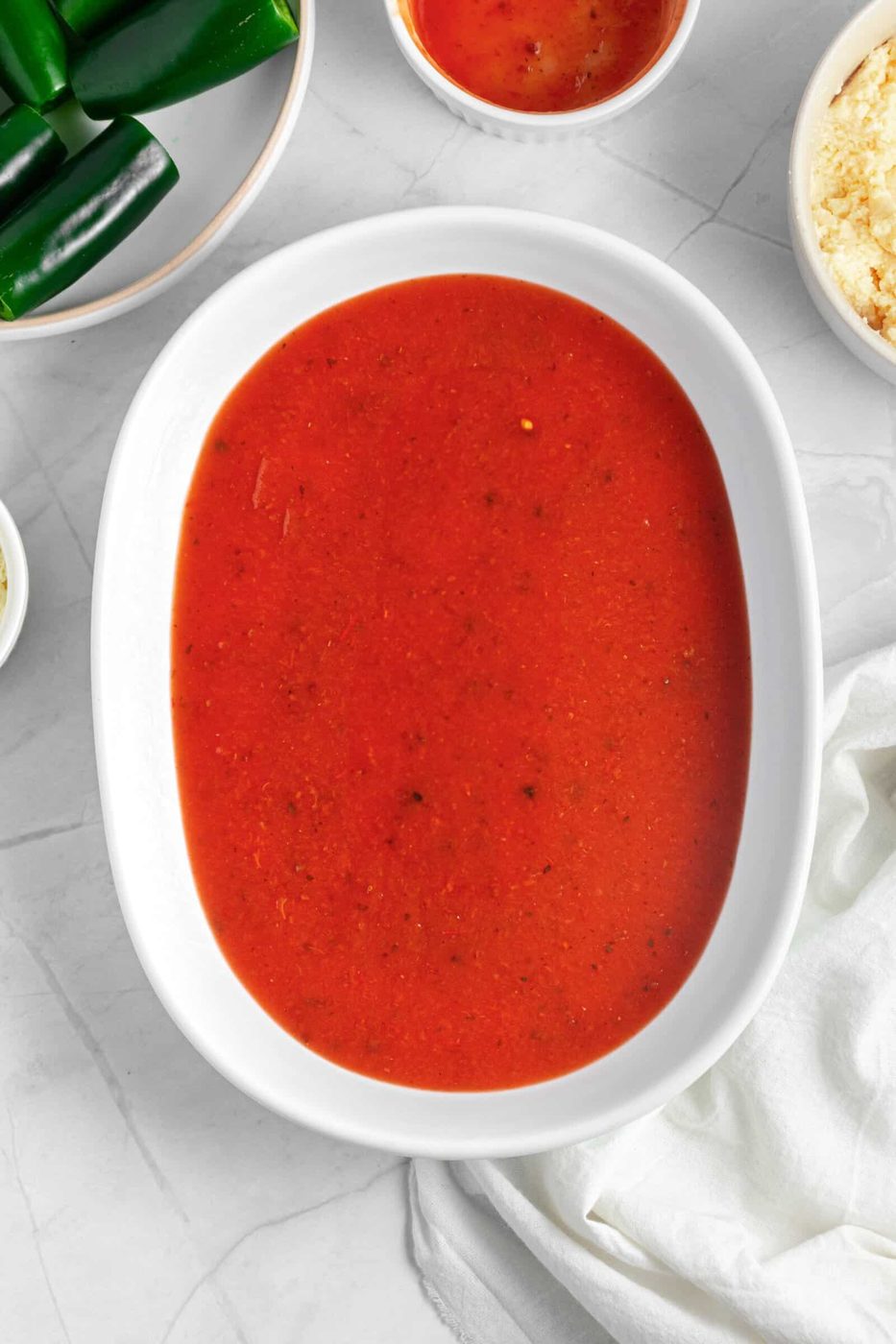 A white oval dish filled with red enchilada sauce on a marble surface. Surrounding the dish are bowls containing sliced green peppers, crumbled cheese, and a small bowl of similar sauce. A white cloth is partially visible on the right.
