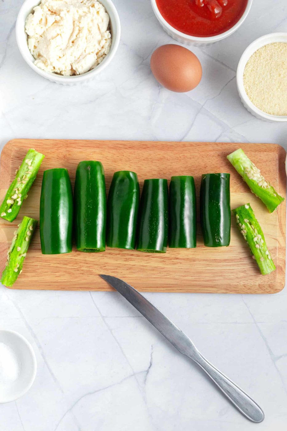 A wooden cutting board with hollowed jalapenos, a knife, and sliced zucchini ends. Surrounding the board are bowls with ricotta cheese, tomato sauce, breadcrumbs, an egg, and a small bowl of salt on a marble countertop.
