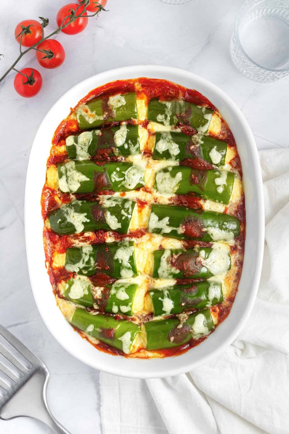 A baked dish of stuffed jalapenos in red tomato sauce, arranged in a white oval dish. The peppers are topped with melted cheese. Nearby are fresh cherry tomatoes on the vine and a glass of water on a marble surface.