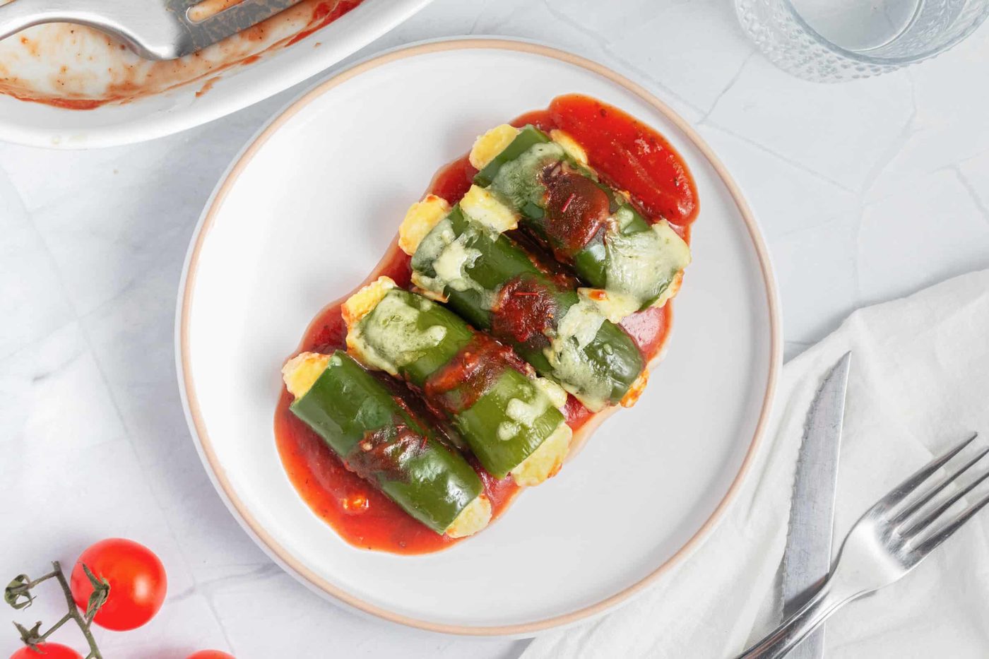 Four green jalapenos are arranged neatly on a white plate, topped with melted cheese and placed on a bed of red sauce. A fork and knife are set nearby, with fresh cherry tomatoes and a glass of water in the background.