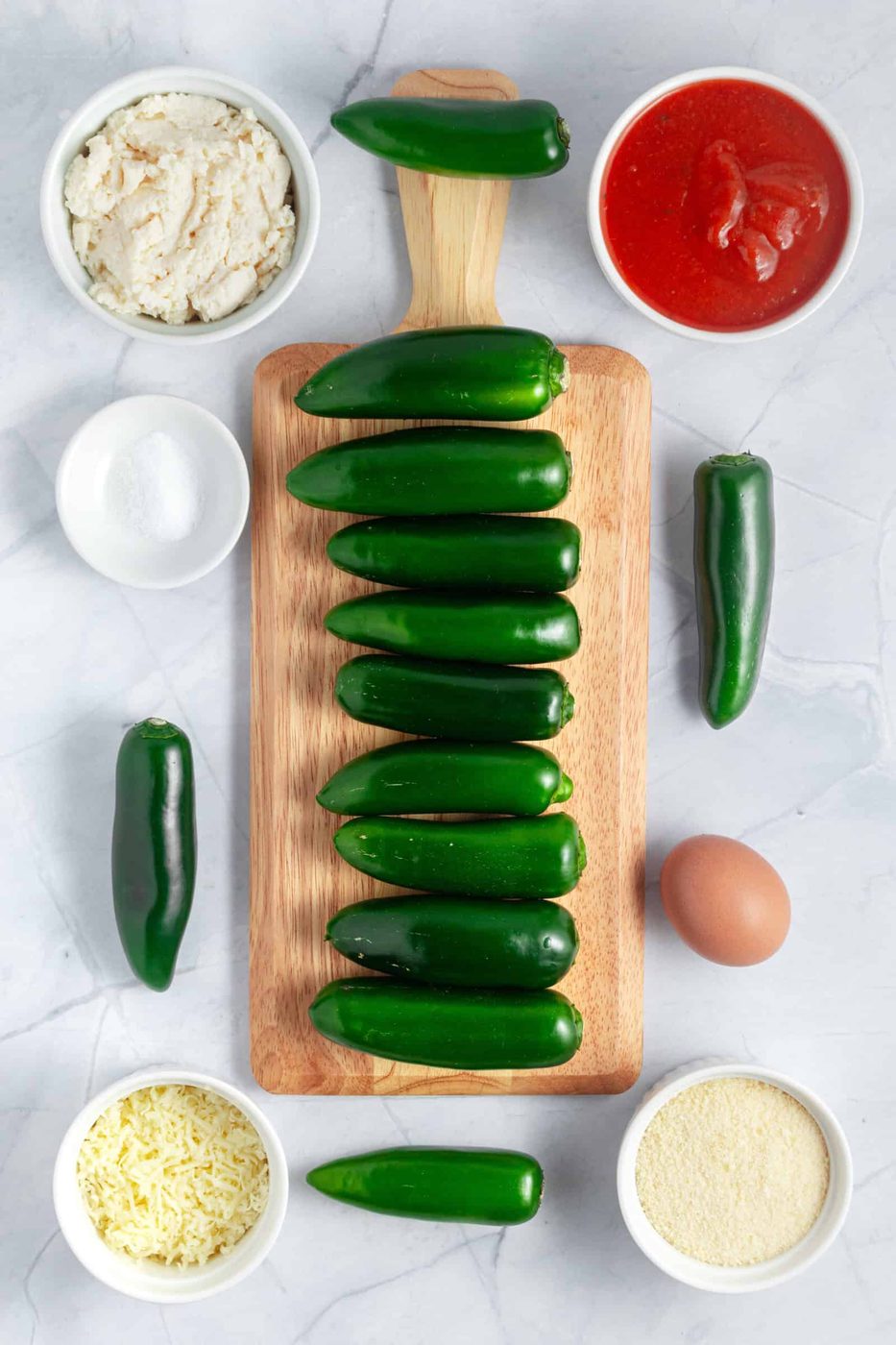 A wooden board with fresh green peppers, surrounded by bowls of ingredients: ricotta cheese, tomato sauce, sugar, shredded cheese, and breadcrumbs. 
