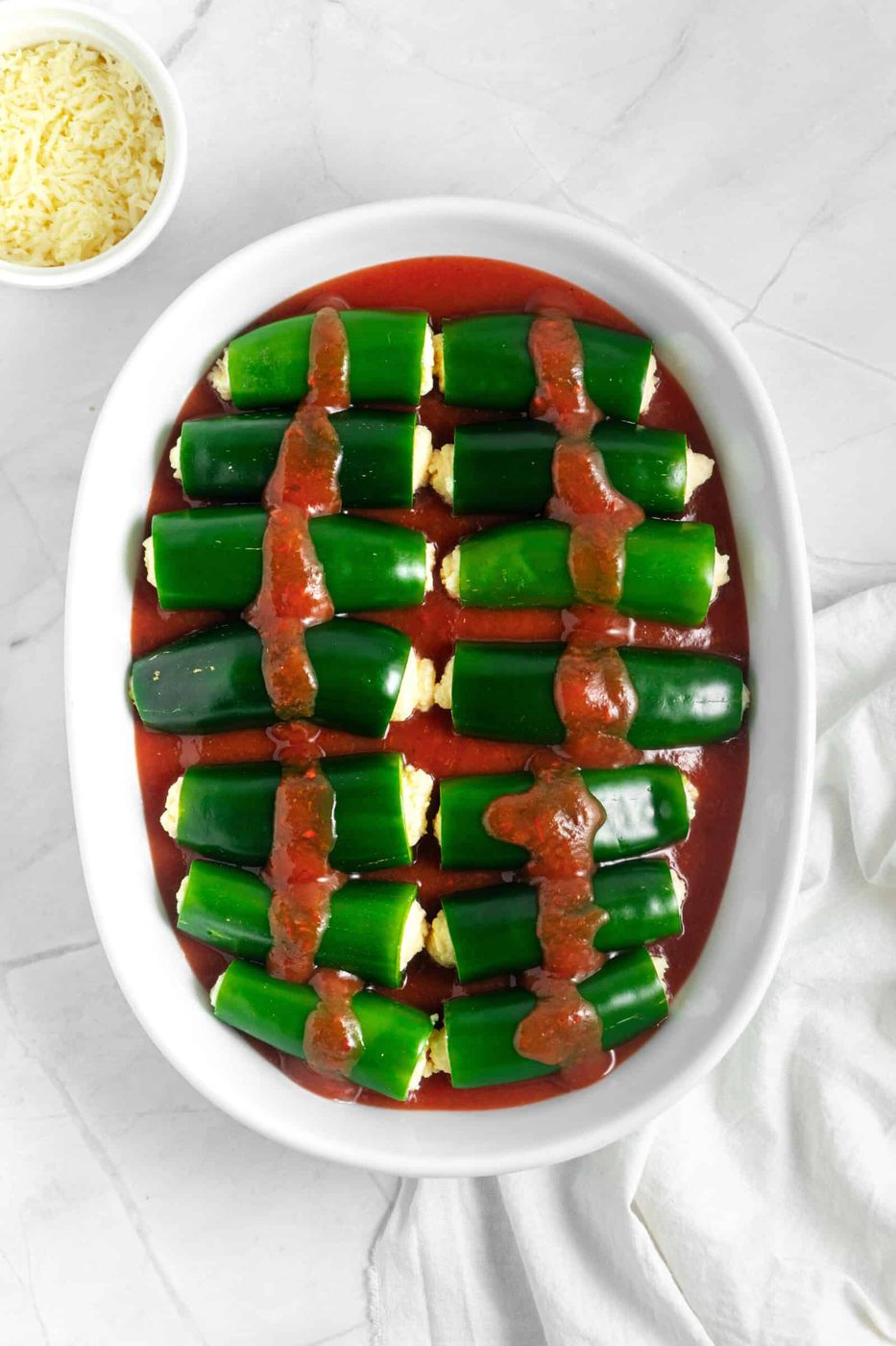 White oval dish with green peppers stuffed and topped with tomato sauce. A small bowl of shredded cheese is in the top left corner. The dish rests on a light marble surface.