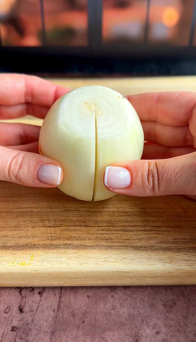 Two hands holding a halved onion with the cut side facing the camera, placed on a wooden cutting board. The background is softly blurred.