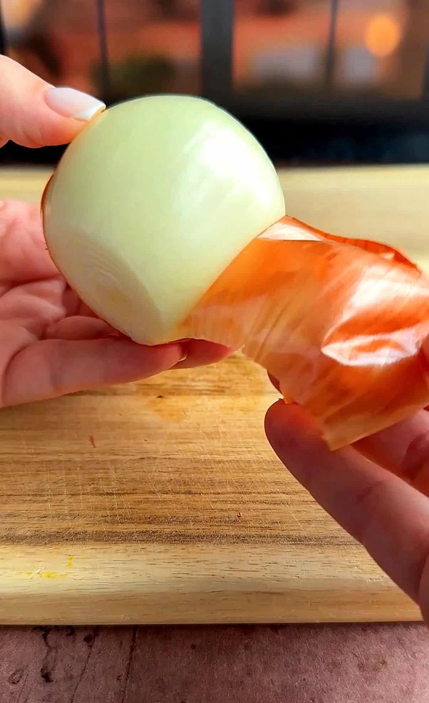 A person peels the skin off a fresh onion over a wooden cutting board. The image focuses on the smooth, glossy surface of the onion, partially covered by papery brown skin.