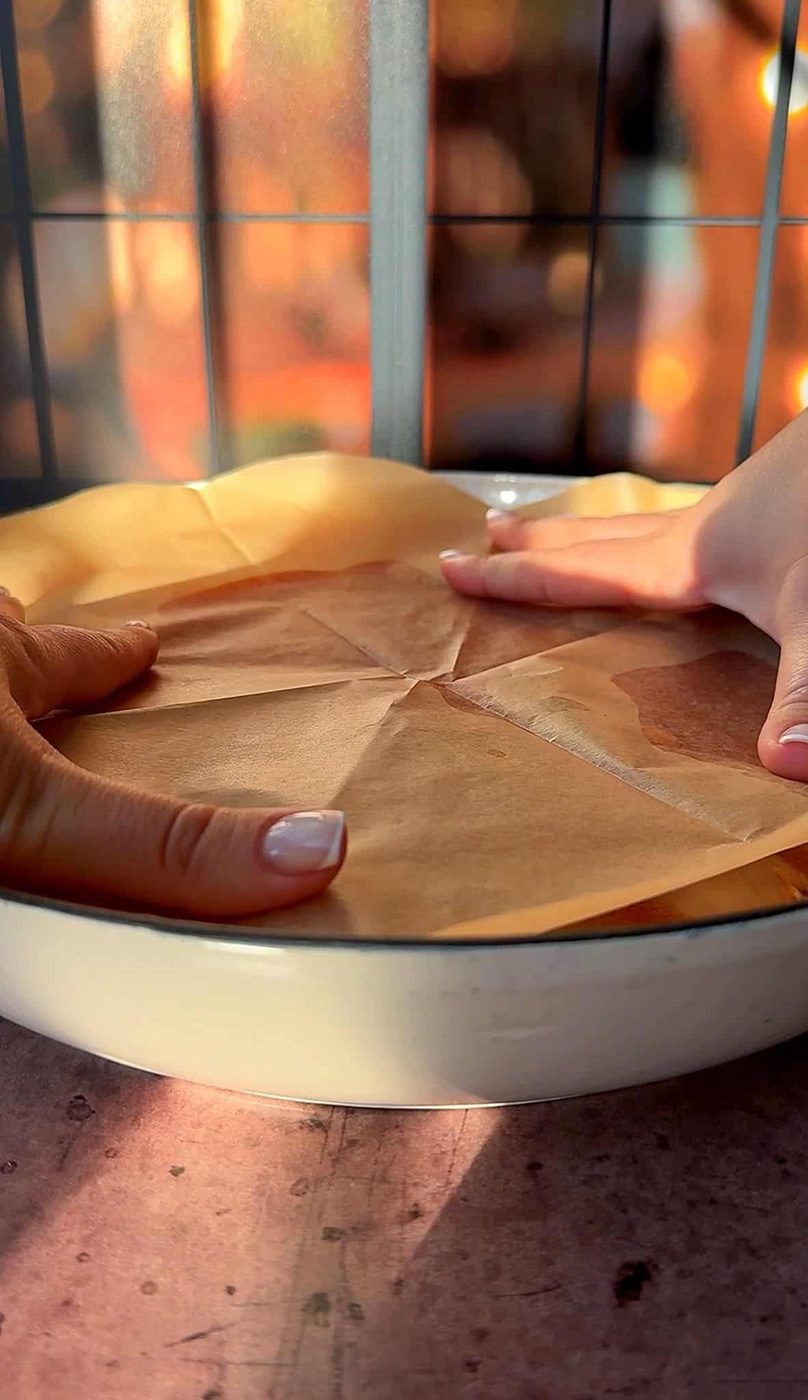 Two hands press parchment paper into a round, white baking dish, on a wooden surface. Soft, warm lighting creates a cozy atmosphere.