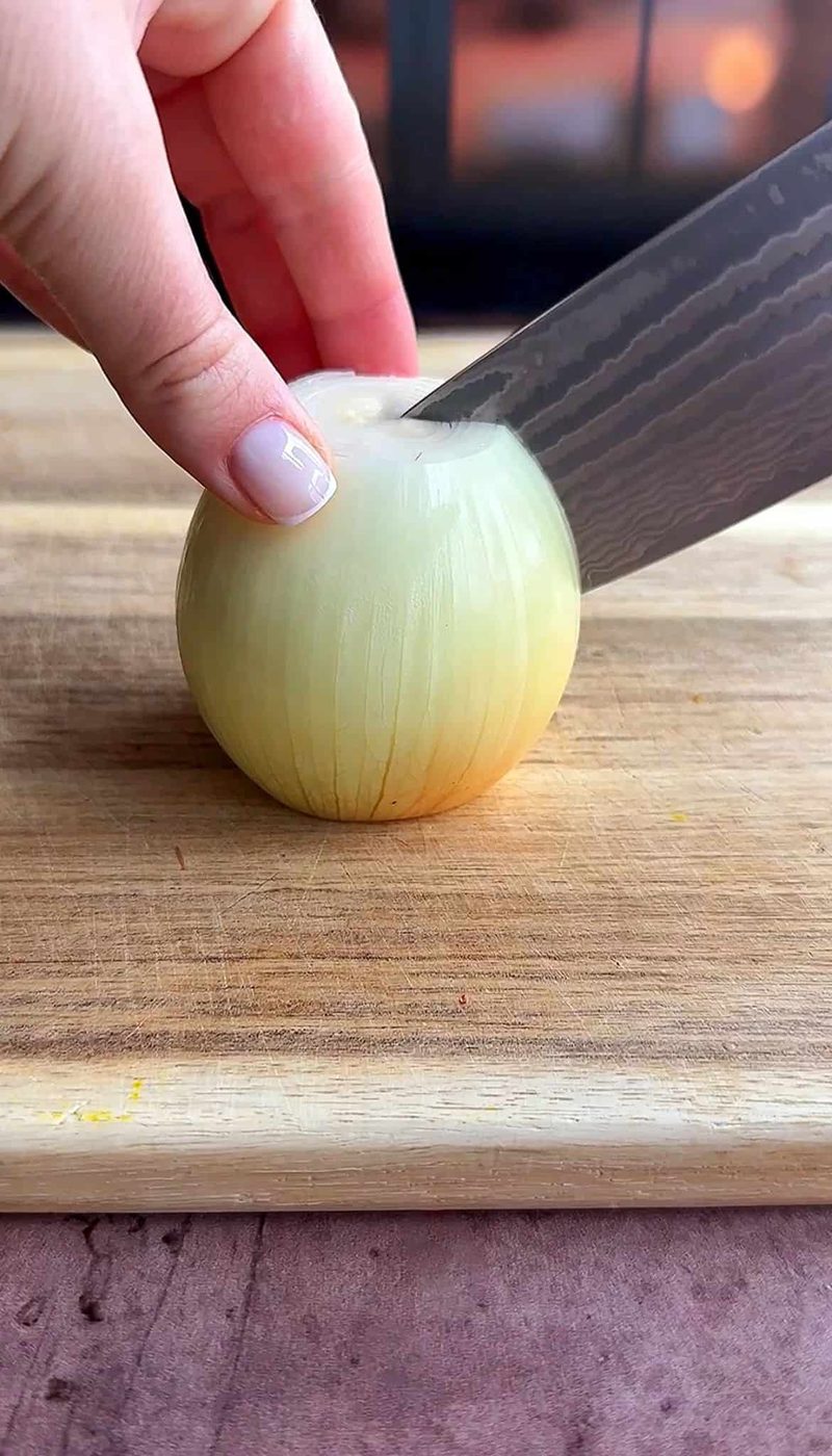 Close-up of a person’s hand holding an onion on a wooden cutting board, while a knife is poised about to slice the onion. The background is softly blurred.