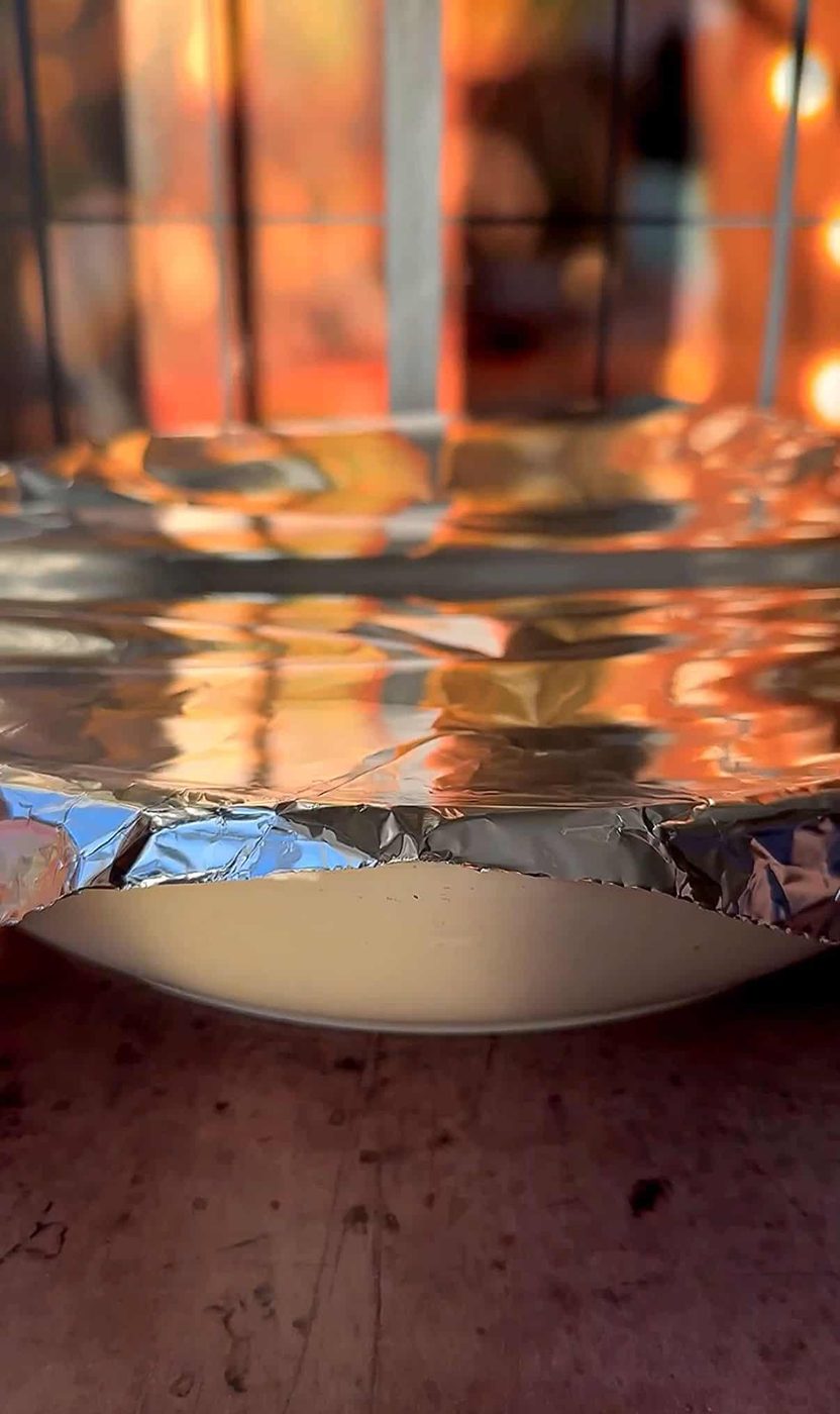 A dish covered with aluminum foil sits on a surface. Warm, out-of-focus autumn colors are visible through a window in the background, creating a cozy atmosphere.
