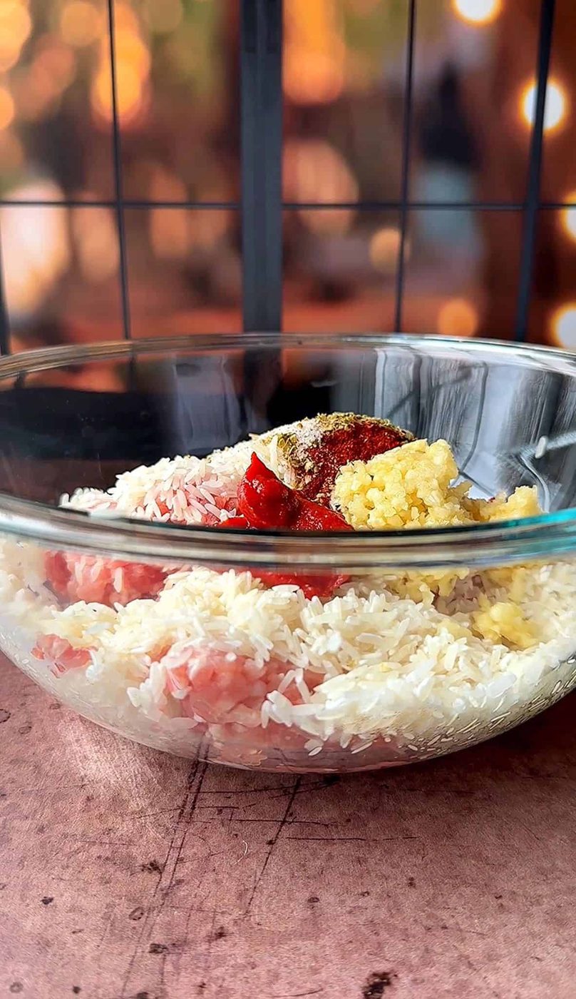 A clear bowl filled with uncooked rice, minced meat, and spices, including a dollop of tomato paste and minced garlic. The background shows a blurred view of warm lights, creating a cozy atmosphere.