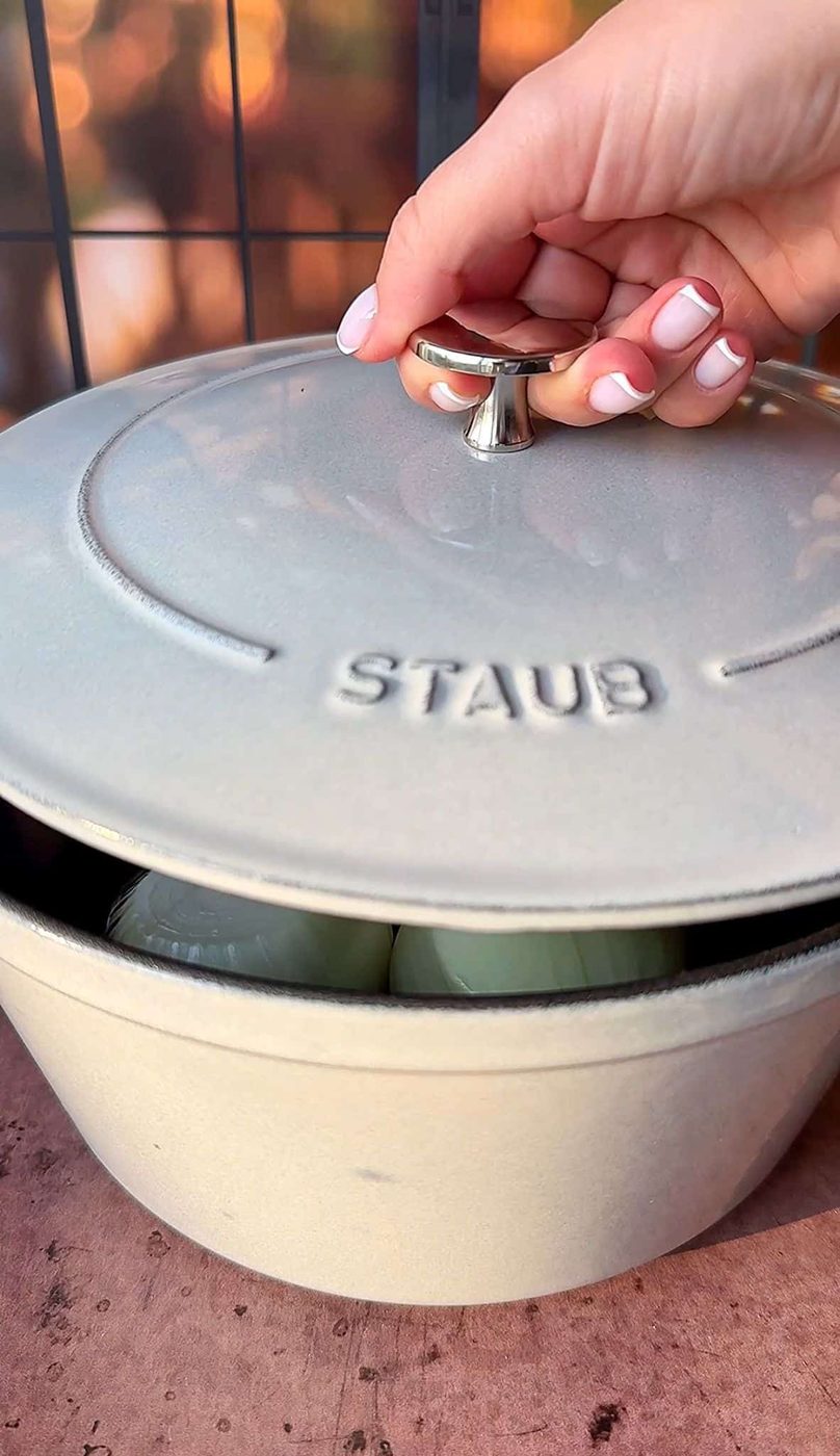 A hand with manicured nails lifts the lid of a Staub Dutch oven, revealing onions inside. The scene is set on a wooden surface with a blurred backdrop, suggesting an outdoor or patio setting.