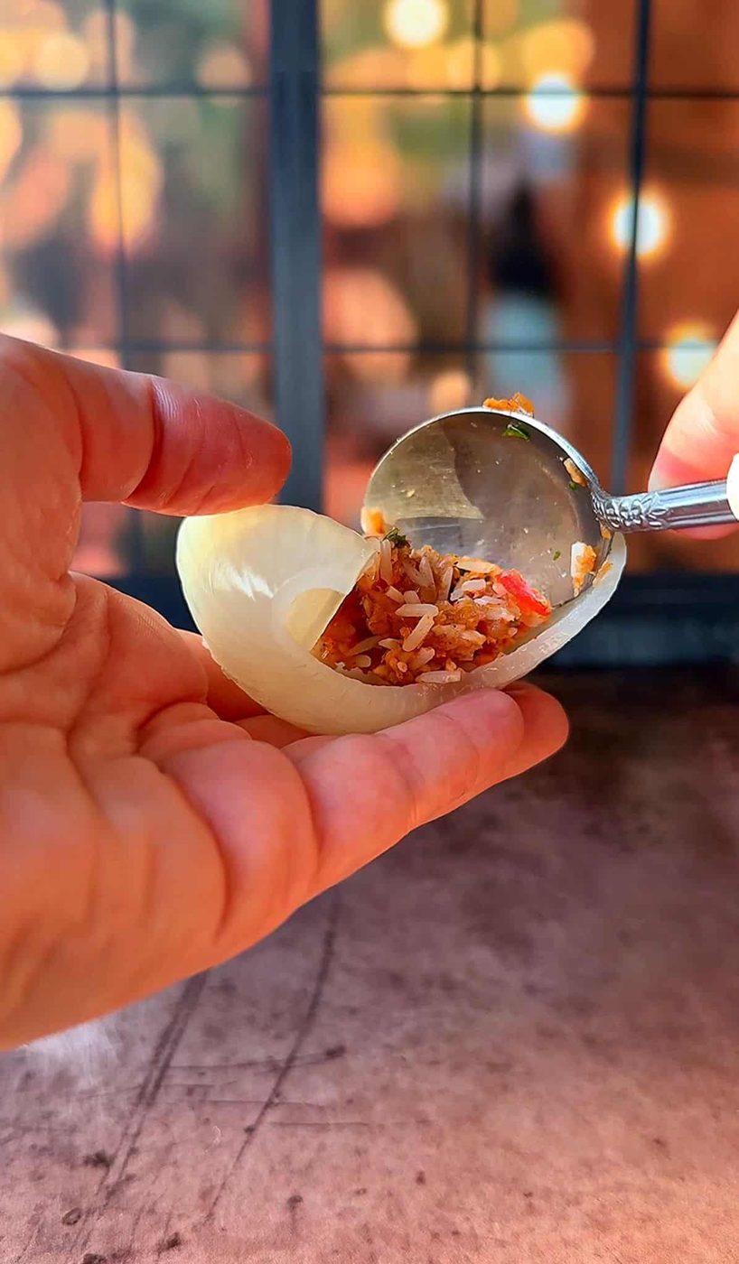 A hand holding a piece of raw onion being filled with a mixture of rice and vegetables using a spoon. The background is softly focused, with warm lighting.