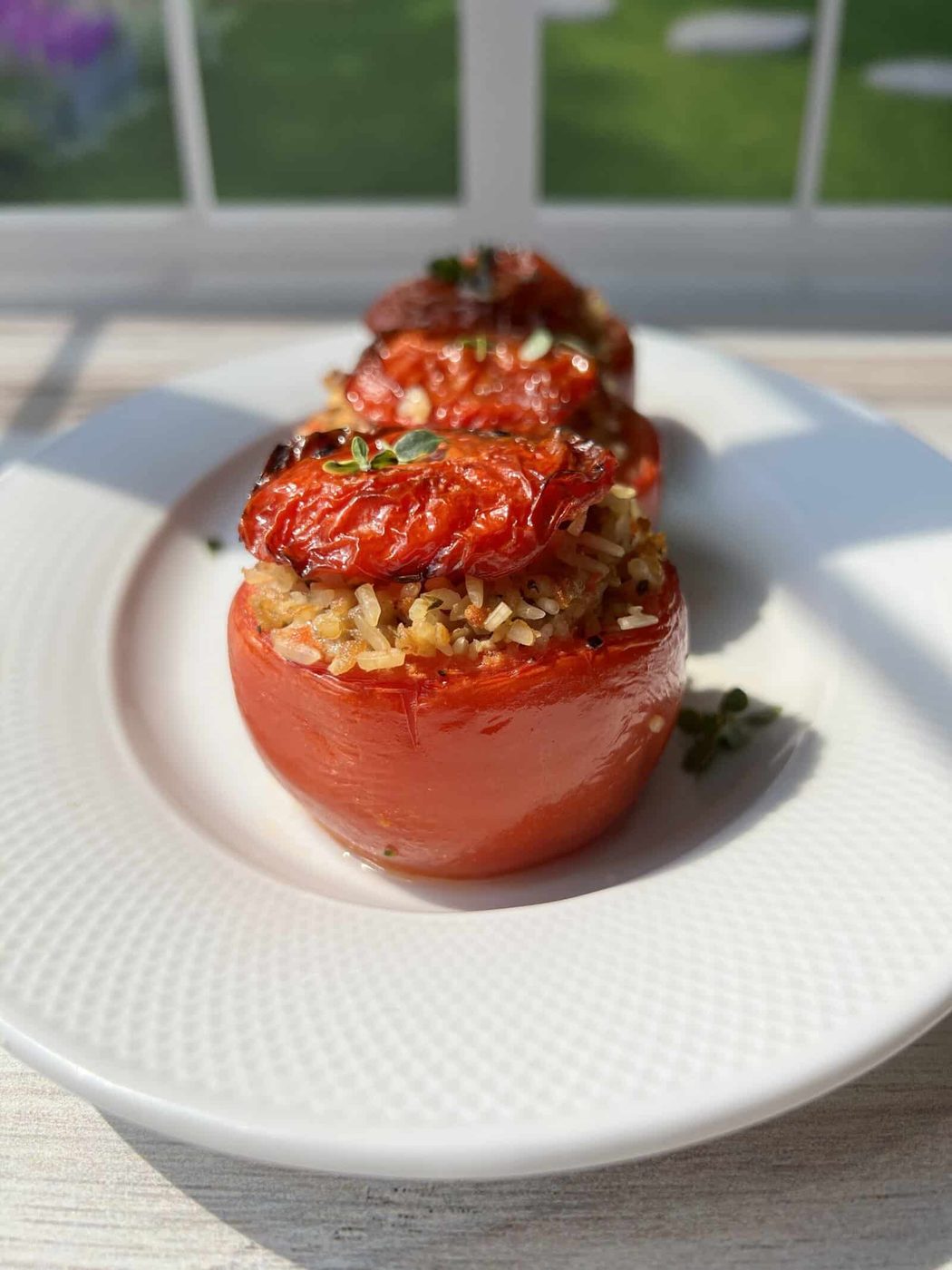 Three stuffed tomatoes filled with rice and herbs, topped with roasted tomato slices and garnished with fresh thyme, served on a white plate with sunlight streaming through a window in the background.