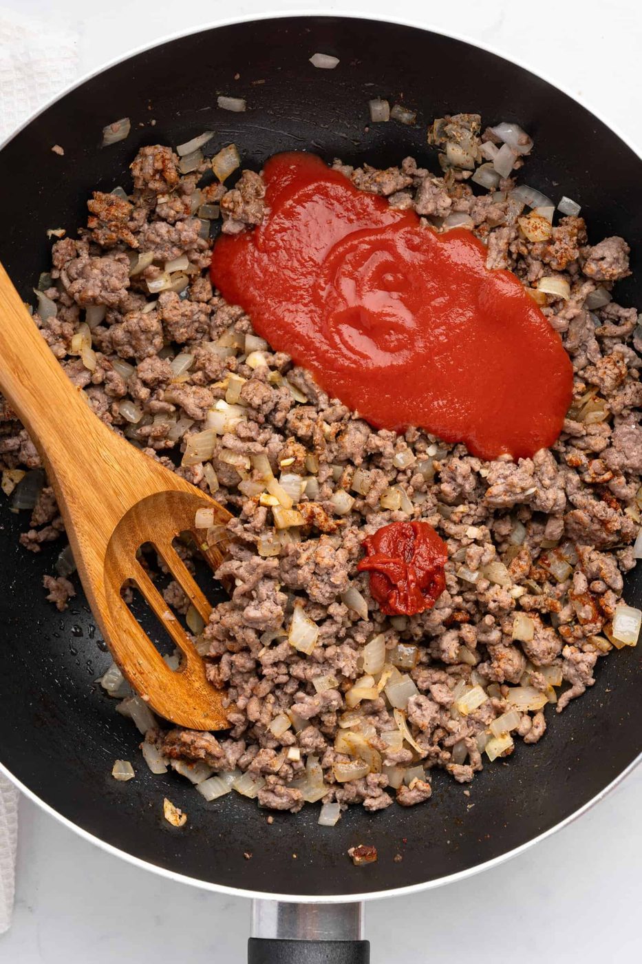 A skillet contains browned ground beef and diced onions, topped with a splash of tomato sauce and a dollop of tomato paste. A wooden spatula rests in the pan, ready to stir the ingredients.