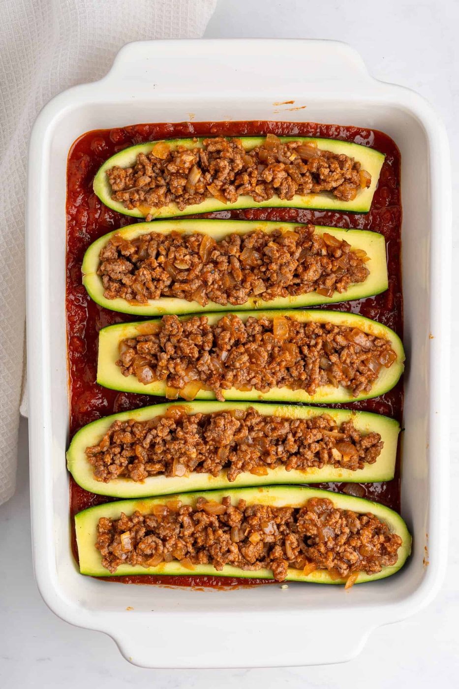 Six zucchini boats filled with seasoned ground meat and vegetables are lined up in a white baking dish. The dish is placed on a light-colored surface. The zucchini boats are cooked in a tomato-based sauce.