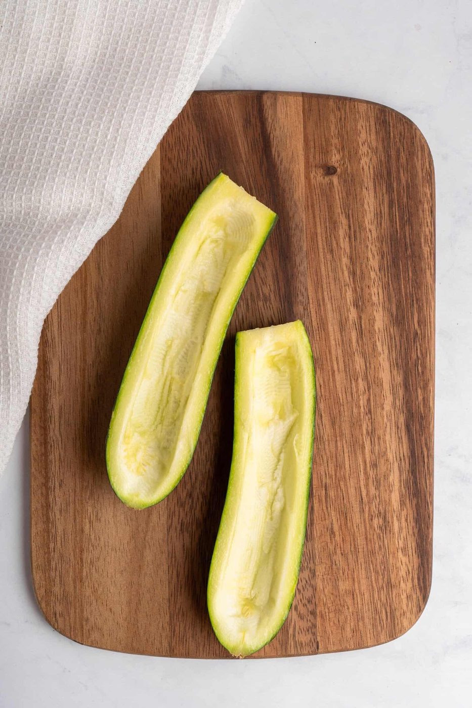 Two zucchini halves with the seeds scooped out, resting on a wooden cutting board. A white cloth partially covers the corner of the board.