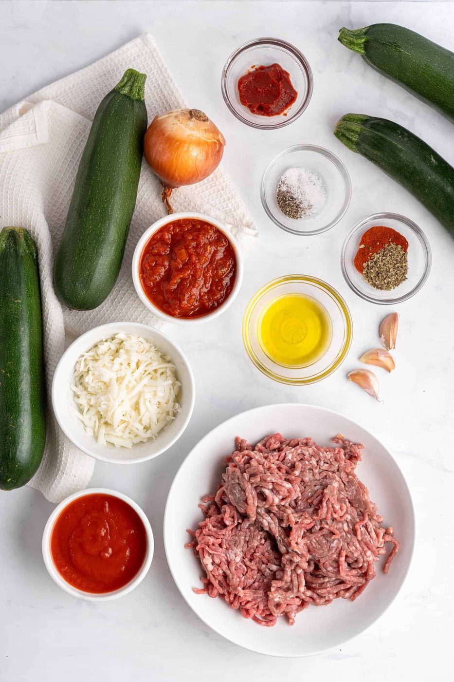 Ingredients for a zucchini recipe arranged on a white surface: three zucchinis, ground meat, olive oil, tomato sauces, cheese, onion, garlic, red pepper flakes, oregano, salt, and pepper.