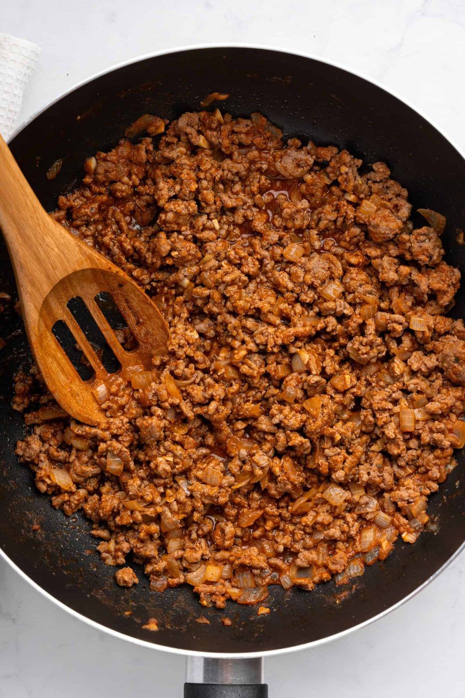 A skillet filled with cooked ground beef mixed with diced onions and spices, with a wooden spatula resting on the side. The mixture appears well-seasoned and is slightly saucy.