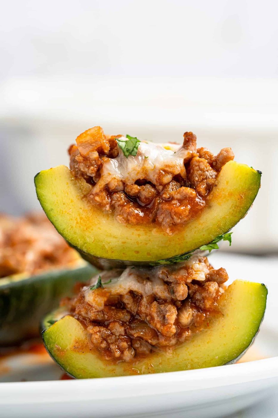 A close-up of two stacked zucchini boats filled with a ground meat mixture, topped with melted cheese and garnished with herbs. The zucchini halves are arranged on a white plate.