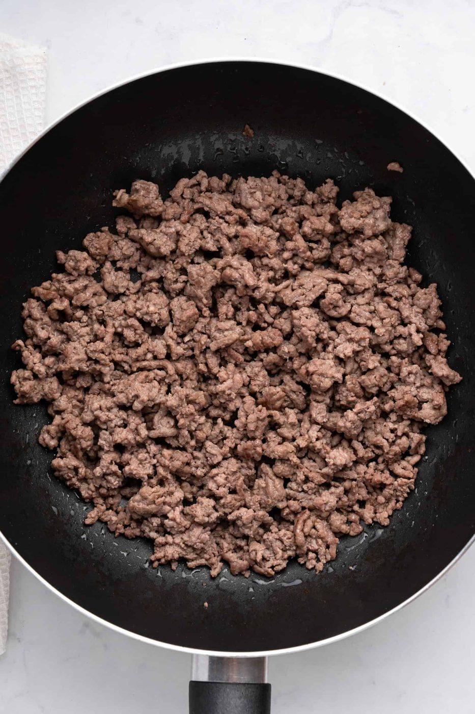A black frying pan filled with cooked, crumbled ground beef sits on a white countertop. The beef is browned and evenly distributed across the pan. A portion of a white cloth is visible on the left side of the image.