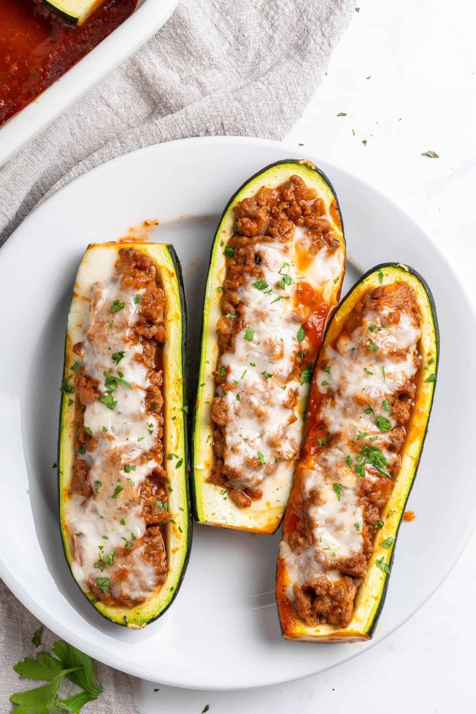 Three zucchini boats filled with a seasoned meat mixture, topped with melted cheese and garnished with parsley, served on a white plate. The dish is placed on a light cloth, with part of a white baking dish visible in the background.