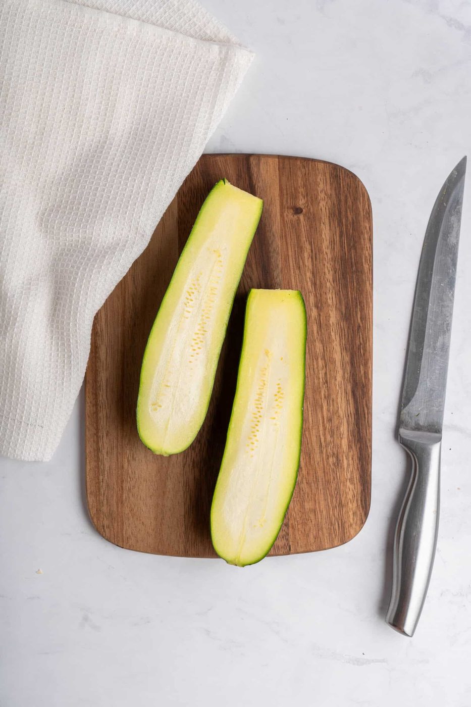 Two zucchini halves are placed on a wooden cutting board. A knife with a metallic handle lies to the right of the board, and a white textured cloth is partially visible to the left on a marble surface.