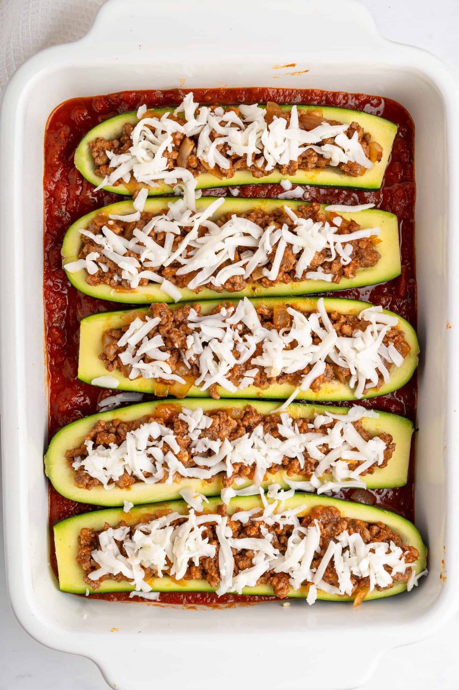 A baking dish filled with zucchini halves stuffed with a mixture of ground meat and topped with shredded cheese. The dish is set in a base of tomato sauce, ready for baking.