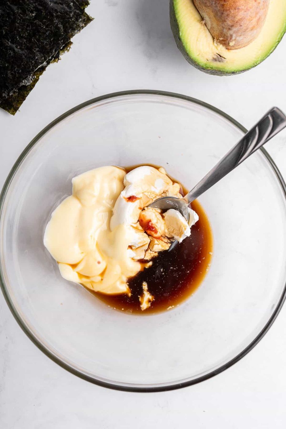 A glass bowl containing mayonnaise, soy sauce, and a small amount of mustard, with a spoon inside. Nearby are a halved avocado and a piece of seaweed on a light-colored surface.