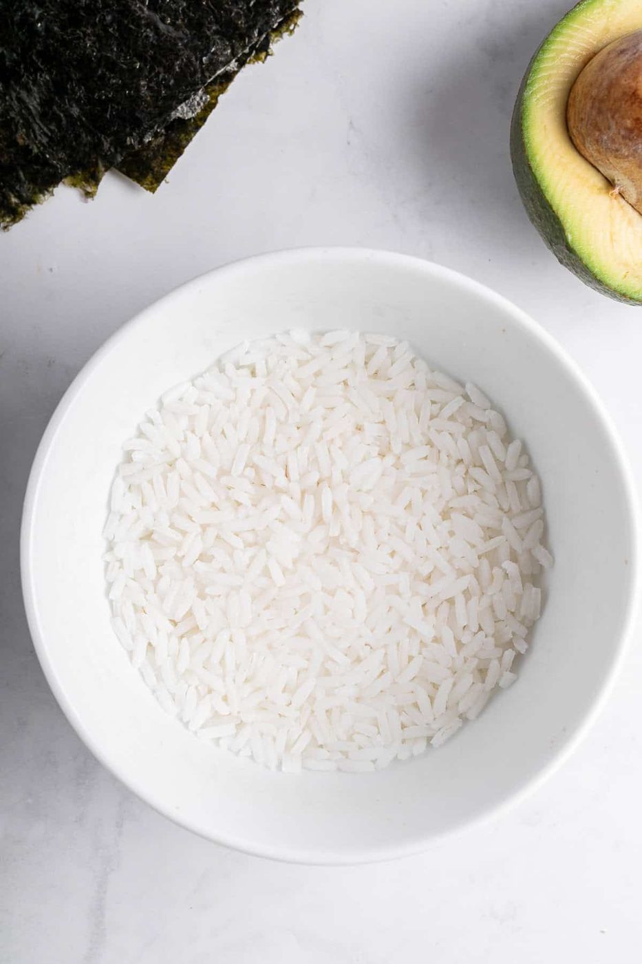 A white bowl filled with uncooked white rice is placed on a marble surface. To the side, there is a halved avocado with the pit visible and a sheet of nori seaweed.