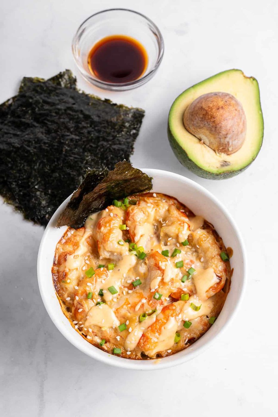 A bowl of creamy baked sushi with shrimp, topped with green onions and sesame seeds. Nori sheets, soy sauce in a small bowl, and a halved avocado are placed nearby on a light surface.