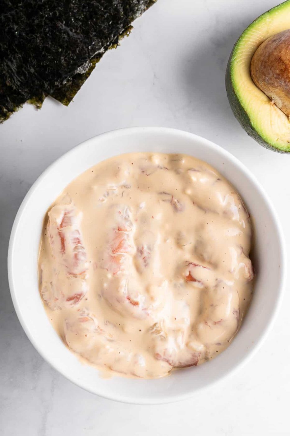 A white bowl filled with a creamy mixture containing chunks of pink seafood, possibly shrimp or crab, is placed on a white marble surface. A partial view of an avocado and sheets of seaweed are visible nearby.