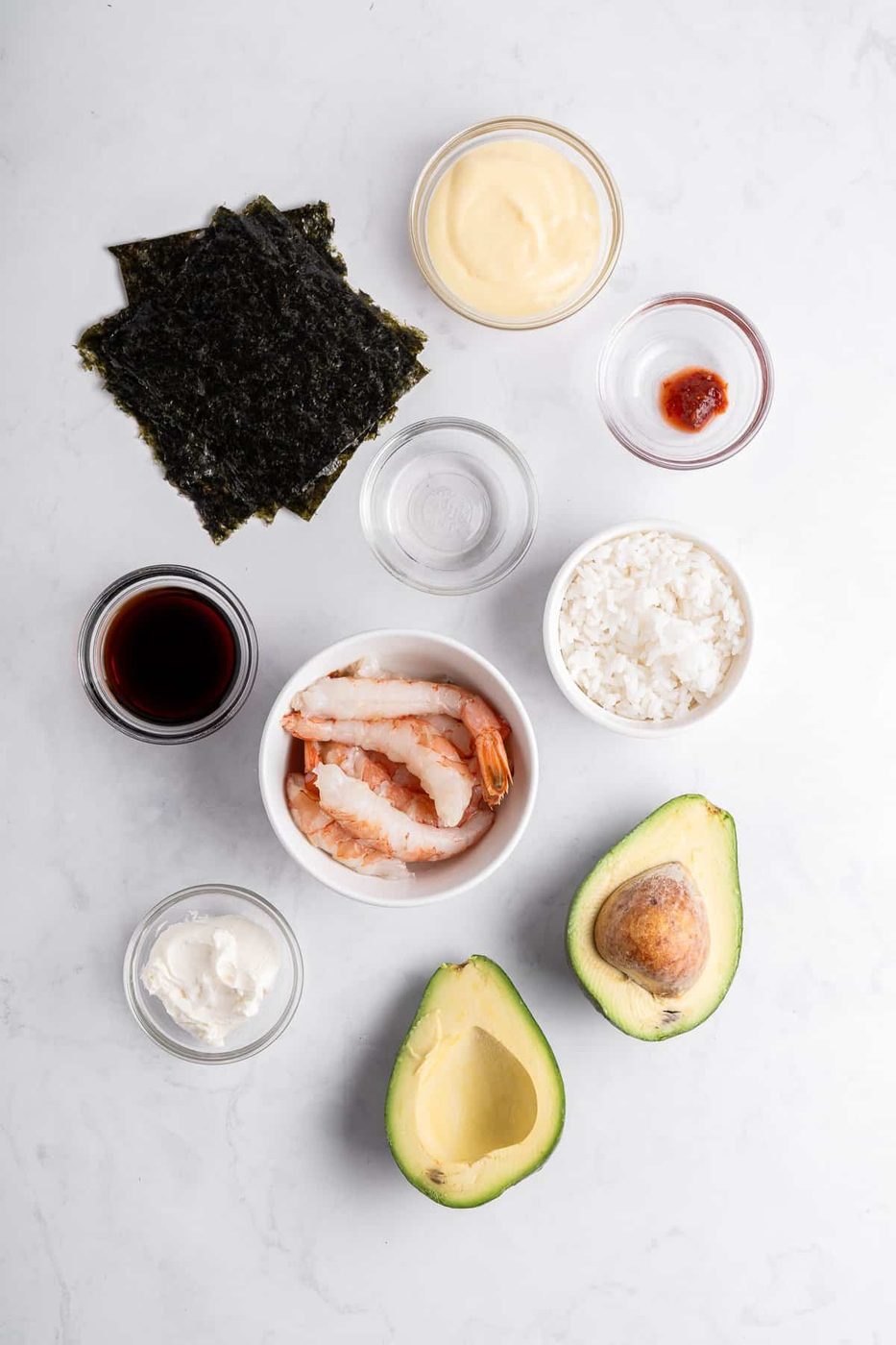 Ingredients for sushi: nori sheets, mayonnaise, soy sauce, shrimp, cooked rice, avocado halves, cream cheese, and a small dish with red chili paste, all arranged on a light marble surface.