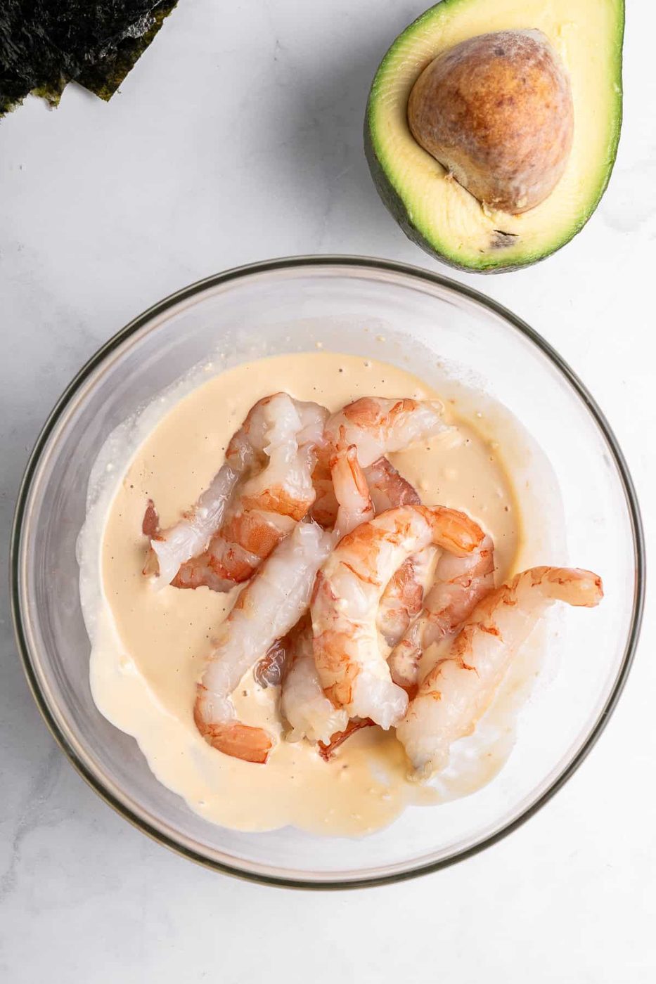 A glass bowl filled with shrimp partially submerged in a creamy sauce. Next to the bowl is a half-avocado with the seed intact. A piece of seaweed is also visible in the top left corner of the image.