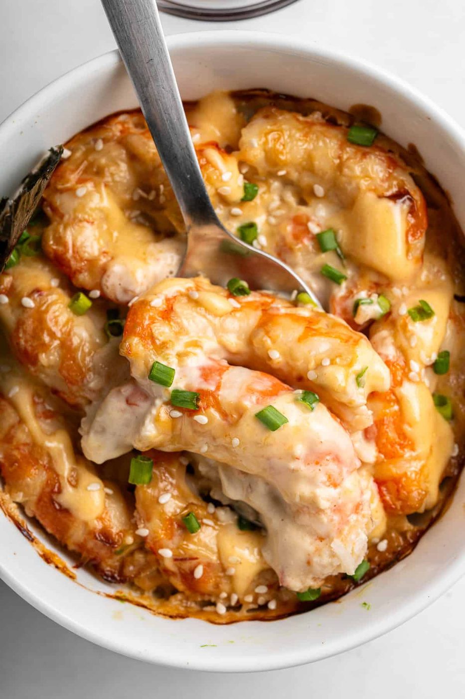 A close-up of a creamy Sushi Bake dish in a white bowl. The shrimp are coated in a cheese sauce, garnished with chopped green onions and sesame seeds. A spoon is partially submerged in the dish, showing the rich texture.