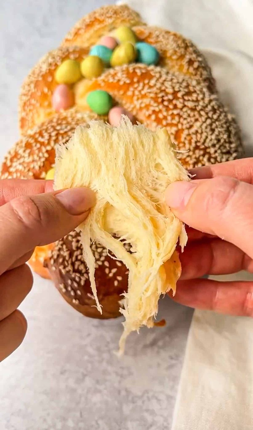 Hands pulling apart a fluffy piece of braided bread topped with colorful candy eggs and sesame seeds, showcasing its soft texture. The background features a gray surface and a white cloth.