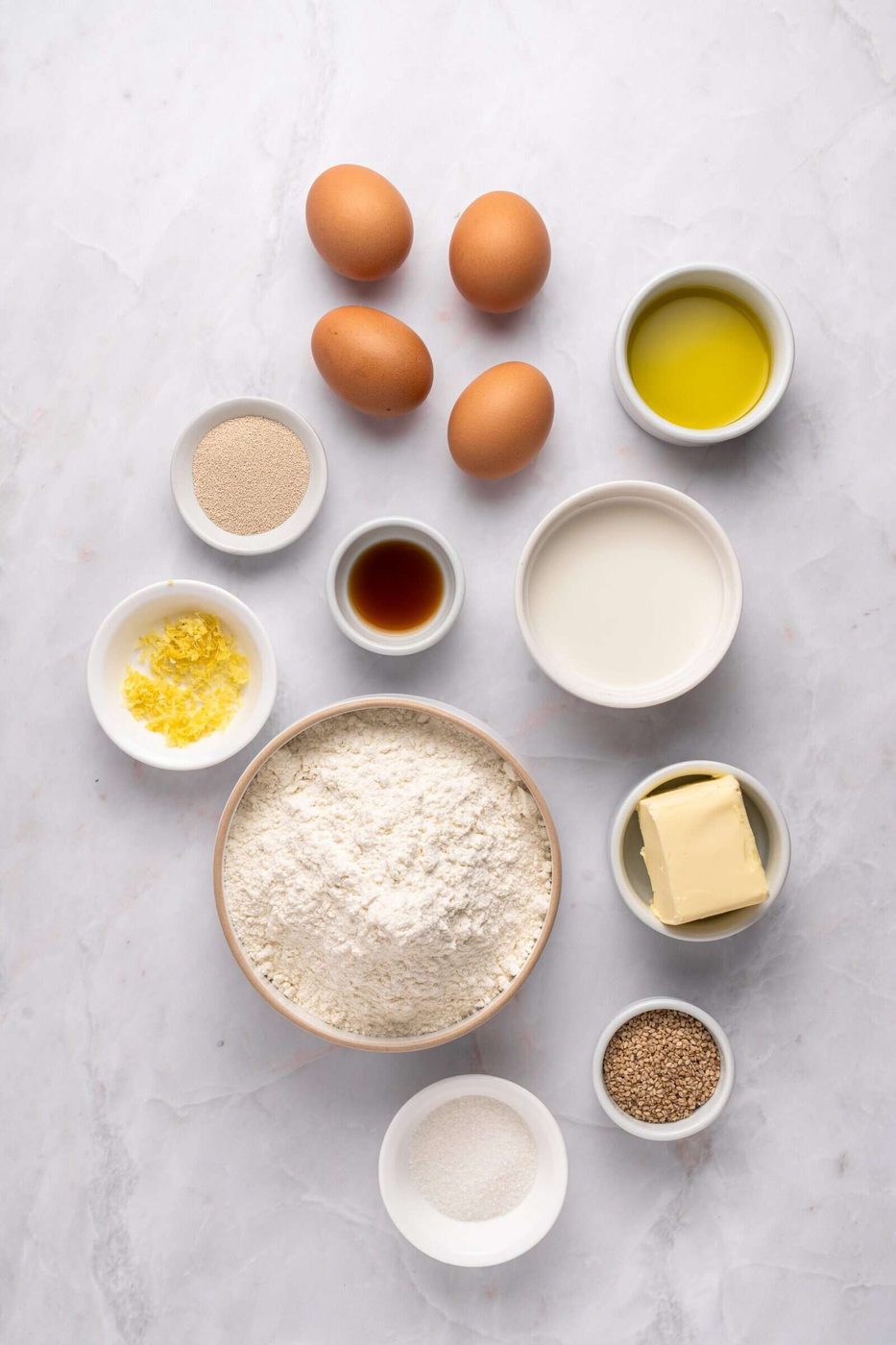 A flat lay of baking ingredients on a white surface, including eggs, olive oil, milk, flour, butter, sugar, vanilla, yeast, lemon zest, and sesame seeds, each in individual bowls or directly on the surface.